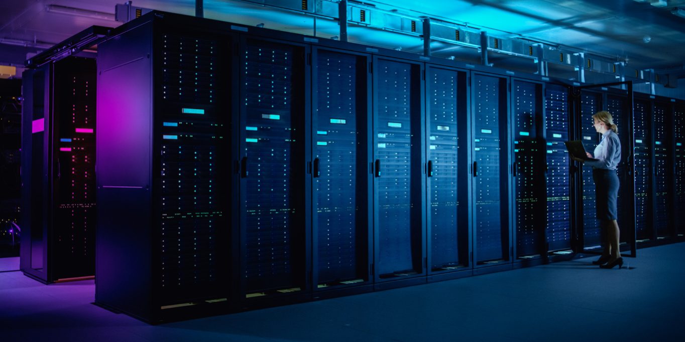 In Data Center: IT Technician Running Maintenance Programme on Laptop, Controls Operational Server Rack Optimal Functioning. Modern High-Tech Telecommunications Operational Data Center in Neon Lights.